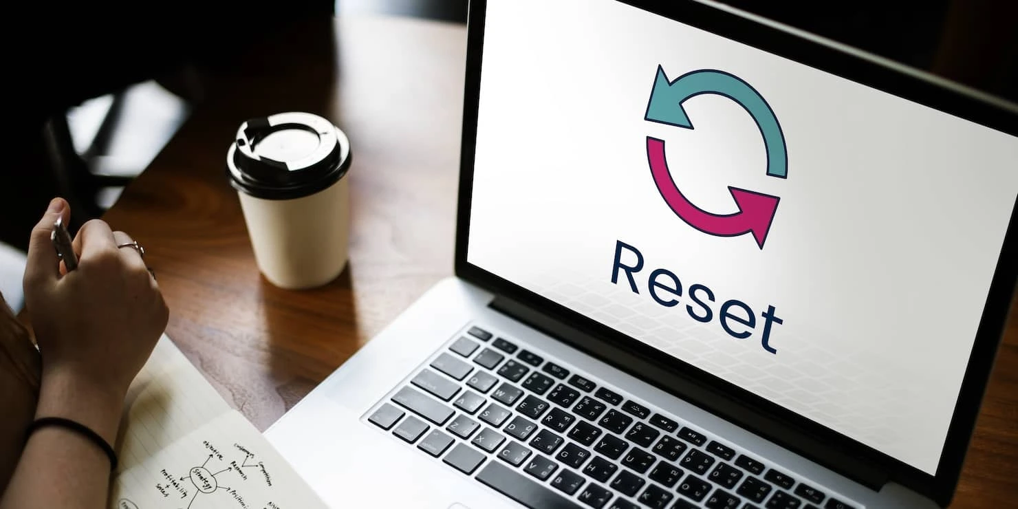 A person's hand is shown next to a coffee cup and a notebook with pen, in front of a laptop screen displaying the word 'Reset' with a circular arrow logo symbolizing refresh or restart.